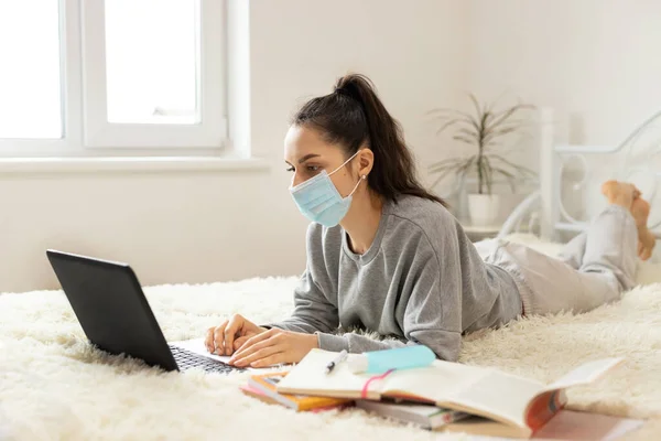 Korumacı tıbbi maske takan genç bir kadın öğrenciler için öğrenim görüyor. Karantinaya alın. Freelancer, evde çalış, online.