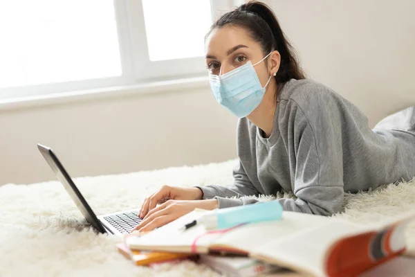 Korumacı tıbbi maske takan genç bir kadın öğrenciler için öğrenim görüyor. Karantinaya alın. Freelancer, evde çalış, online.