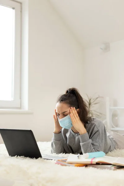 woman girl in protective mask, with a high fever and headache, works with a laptop during quarantine,
