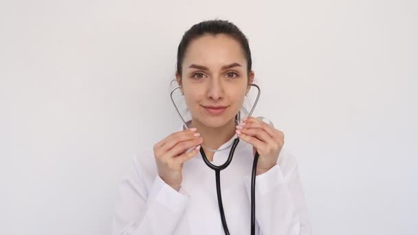 Primer plano joven sonriente doctor mujer chica en un blanco médico abrigo, pone un estetoscopio en sus oídos, mira a la cámara — Vídeo de stock