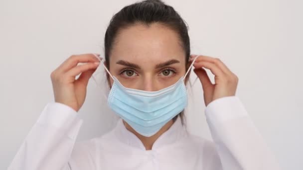Närbild ung leende läkare sjuksköterska kvinna flicka i en vit medicinsk rock, sätter på en medicinsk skyddsmask, tittar på kameran — Stockvideo
