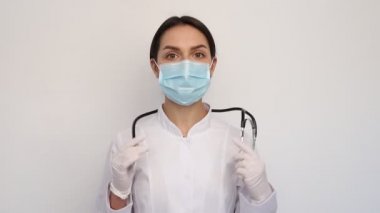 Beyaz tıbbi eldiven ve koruyucu maske takan kadın doktor boynuna steteskop takıyor.