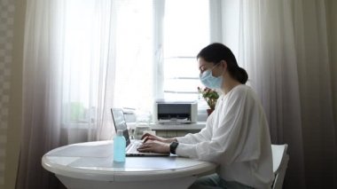 Ciddi, koruyucu maskeli, evde dizüstü bilgisayarla e-posta yazan, bilgisayarla internetten ders çalışan, sandalyeye oturmuş, karantina kovid 19 'daki bir kız.
