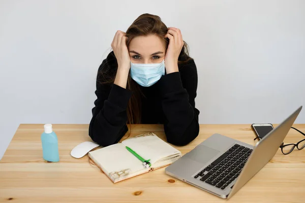 woman girl wearing a protective mask sits at a table, with a high fever and headache, works with a laptop during quarantine, the concept of health and coronavirus, on the table sanitizer for hands