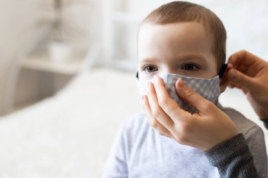 Tıbbi koruyucu maskeli küçük bir çocuk. Anne, bebek steril tıbbi maskesini takıyor. Çocuk, yüz maskesi takıyor, karantinada virüs, salgın ve salgın hastalıklardan korunuyor.