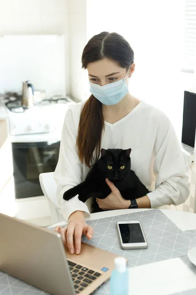 woman girl in a medical mask works on a laptop, holds a black cat in her arms, social distance during the epidemic coronavirus covid-19, work from home, online education, hand sanitizer on the table