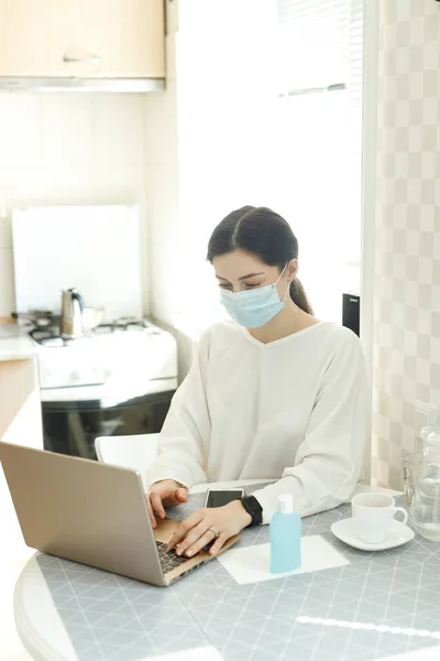 woman girl in medical mask works on laptop computer, social distance during epidemic of influenza virus coronavirus covid-19, work at home, online training, health concept, hand sanitizer on the table