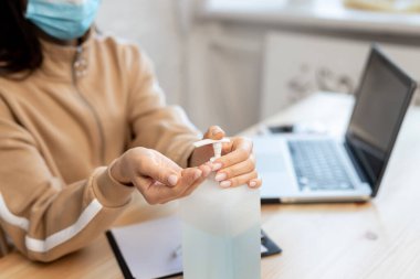 Koruyucu tıbbi maske takan genç bir kızı giydirip dezenfektan dağıtıyor ve COVID-19 karantinası sırasında masada dizüstü bilgisayarla çalışıyor. Coronavirus koruması.