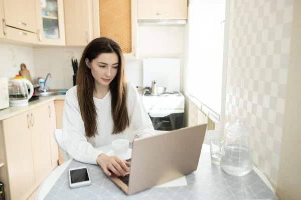 The girl writing on laptop in kitchen. Young businesswoman work with laptop at home. Remote working concept. The young girl freelancer copywrite from home.