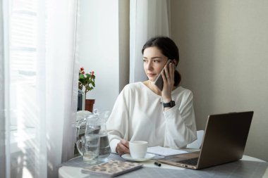 İş kadını danışmanlık yapıyor ve evde kahve içiyor. Uzaktan kumandalı yetenekli yönetici dizüstü bilgisayarda e-posta okuyor. Uzaktan çalışma ve telefon danışma konsepti. Pencerede Göster
