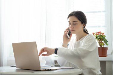 Genç iş kadını telefonla danışmanlık yapıyor ve evdeki akıllı saati kontrol ediyor. Uzaktan çalışma ve telefon danışma kavramı