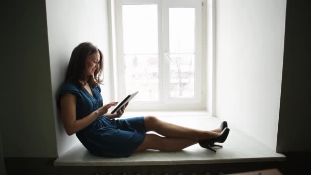 Prores. Menina em um vestido azul está sentado com um tablet na janela — Vídeo de Stock