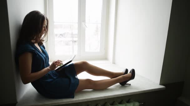 Prores. Chica joven en un vestido azul está sentado con una tableta en la ventana — Vídeos de Stock