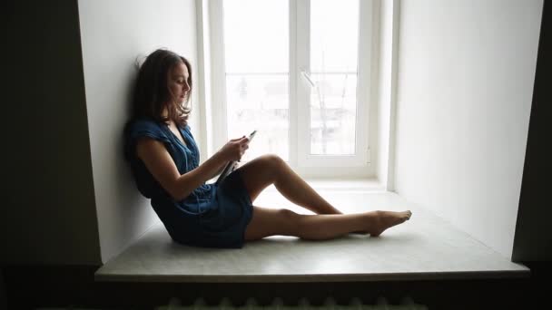 Prore. Giovane ragazza in un abito blu è seduto con un tablet sulla finestra — Video Stock