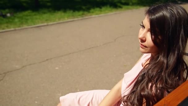 一个穿着粉红色连衣裙的年轻女孩坐在公园的长凳上 — 图库视频影像