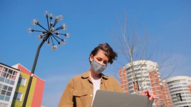 Kahverengi elbiseli ve maskeli genç bir kadın parktaki arka planda dizüstü bilgisayarla uzaktan çalışıyor.