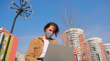 Kahverengi elbiseli ve maskeli duygusal genç bir kadın dışarıda dizüstü bilgisayarla şehrin arka planına karşı çalışıyor.