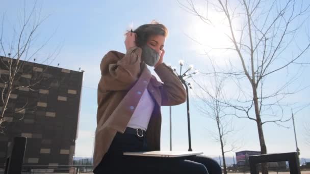 穿着棕色衣服的年轻妇女在城市公园里戴上医疗面具 然后才在笔记本电脑上工作 — 图库视频影像