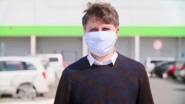 Yüzünden tıbbi bir maske çıkaran bir adamın yakın plan portresi bir alışveriş merkezi ve araba parkının arka planında.