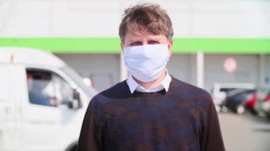 Yüzünde tıbbi maske olan bir adamın yakın plan portresi bir alışveriş merkezi ve araba parkının arka planına karşı.