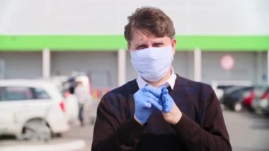 Yüzünde tıbbi maske olan bir adamın yakın plan portresi bir alışveriş merkezi ve araba parkının arka planına karşı.