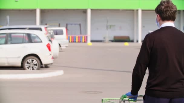 一个头戴医疗面罩 戴着蓝色手套的男人开着购物车沿着购物中心前面的停车场走着 — 图库视频影像