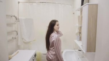 Pembe gecelikli uzun siyah saçlı genç kız banyoda dişlerini fırçalarken ve yıkarken eğleniyor.