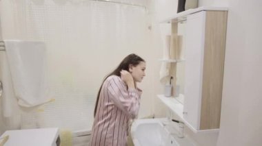 Pembe gecelikli uzun siyah saçlı genç kız banyoda dişlerini fırçalarken ve yıkarken eğleniyor.