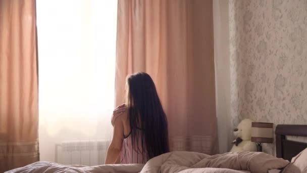 一个穿着浅色睡衣的黑头发少女在卧室的一张大床上醒来 打开窗户上的窗帘 — 图库视频影像