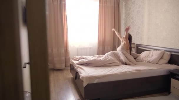 一个穿着浅色睡衣的黑头发少女在卧室的一张大床上醒来 打开窗户上的窗帘 — 图库视频影像