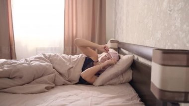 Uzun saçlı, açık siyah gecelikli, uyku bandı takan neşeli, genç bir esmer büyük bir yatakta, bej bir yatak odasında uyanır.