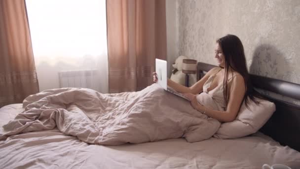年轻活泼的黑发女孩穿着浅色睡衣 躺在卧室的一张大床上 在笔记本电脑上工作 — 图库视频影像