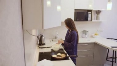 Uzun siyah saçlı ve mavi elbiseli bir kız apartman mutfağındaki tavada sağlıklı bir kahvaltı hazırlar.