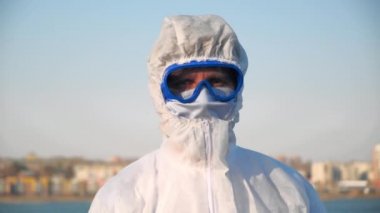 Koruyucu kıyafetli, maskeli ve gözlüklü bir erkek doktorun geniş bir portresi. Enfeksiyon salgını sırasında açık havada temas olmayan termometrenin sıcaklığını ölçen bir virüs taşıyor.