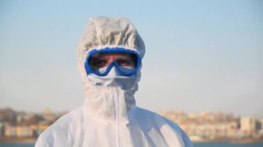 Koruyucu kıyafetli, maskeli ve gözlüklü bir erkek doktorun geniş bir portresi. Enfeksiyon salgını sırasında açık havada temas olmayan termometrenin sıcaklığını ölçen bir virüs taşıyor.
