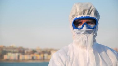 Koruyucu kıyafetli, maskeli ve gözlüklü bir erkek doktorun geniş bir portresi. Enfeksiyon salgını sırasında açık havada temas olmayan termometrenin sıcaklığını ölçen bir virüs taşıyor.