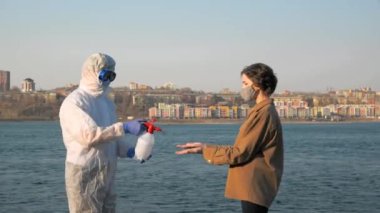 Koruyucu giysili, maskeli ve gözlüklü bir doktor virüs salgını sırasında genç bir kadının ellerini şehrin ve nehrin arka planına dezenfekte ediyor.