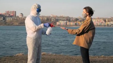 Koruyucu giysili, maskeli ve gözlüklü bir doktor virüs salgını sırasında genç bir kadının ellerini şehrin ve nehrin arka planına dezenfekte ediyor.