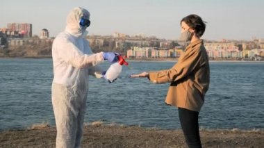 Koruyucu giysili, maskeli ve gözlüklü bir doktor virüs salgını sırasında genç bir kadının ellerini şehrin ve nehrin arka planına dezenfekte ediyor.