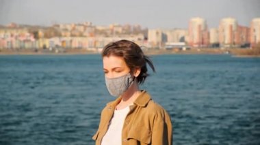 Kahverengi elbiseli ve yüzünde tıbbi maske olan genç bir kadının portresi şehrin ve nehrin arka planında.