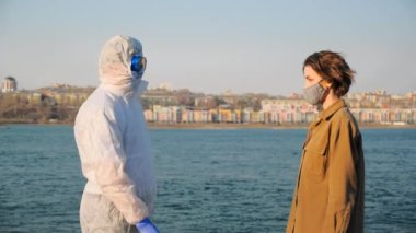 Virüs salgını sırasında koruyucu giysili, maskeli ve gözlüklü bir doktor şehrin ve nehrin arka planında genç bir kadının temassız termometresinin sıcaklığını ölçer.