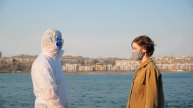 Virüs salgını sırasında koruyucu giysili, maskeli ve gözlüklü bir doktor şehrin ve nehrin arka planında genç bir kadının temassız termometresinin sıcaklığını ölçer.