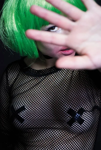unknown woman in black transparent dress from the grid green wig and taped nipples covers her face with hand