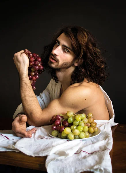 Foto hermoso chico desnudo con pelo largo y rizado con cortinas blancas con uvas en la mano en la mesa — Foto de Stock