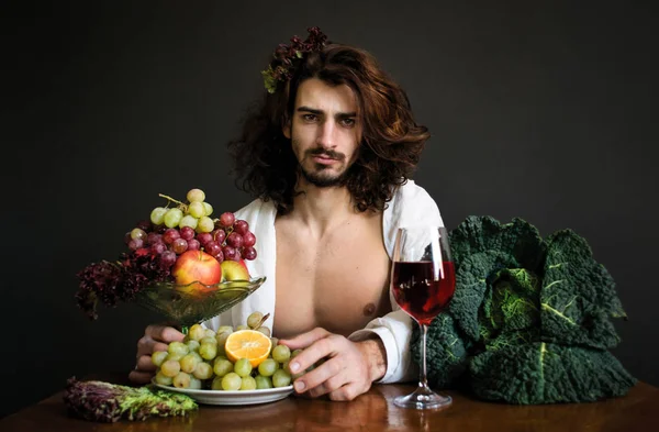 Foto chico guapo medio desnudo con el pelo rizado largo con un vaso de bebida roja y un plato de uvas y frutas en la mesa — Foto de Stock