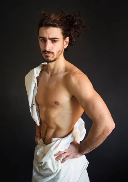 photo half naked handsome guy with long curly hair with white drapery is hand on waist