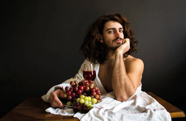 Foto de chico guapo con el pelo largo y rizado en la mesa con un vaso de bebida roja y un plato de uvas — Foto de Stock