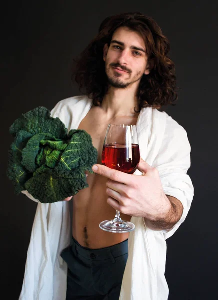 Фото красивый парень в белой драпировке держа свежую зеленую капусту и бокал вина в руке — стоковое фото