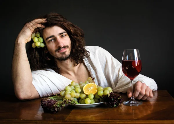 Chico rizado medio desnudo en la mesa con un plato de uvas y una copa de vino foto — Foto de Stock