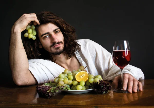 Foto chico rizado medio desnudo en la mesa con un plato de uvas y una copa de vino — Foto de Stock